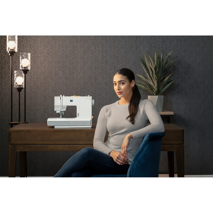 Woman sitting in front of a sewing machine on a wooden table with a dark wall and plant in the background.