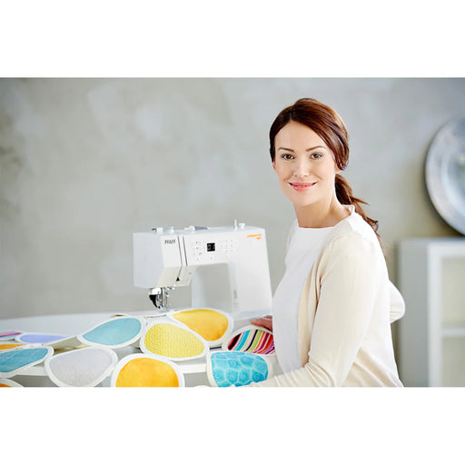 Woman sitting at a sewing machine with colorful fabric samples in a bright room.