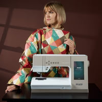 Woman wearing a colorful apron sitting next to a PFAFF sewing machine on a dark surface with a brown background.