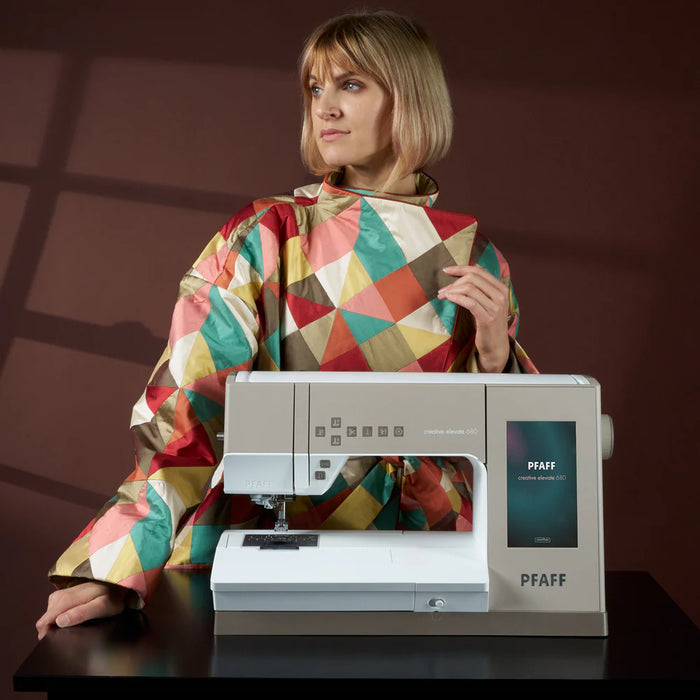 Woman wearing a colorful apron sitting next to a PFAFF sewing machine on a dark surface with a brown background.
