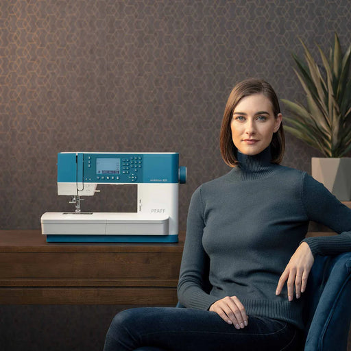 Woman sitting next to a Brother sewing machine on a wooden table with a decorative wall and plant in the background.